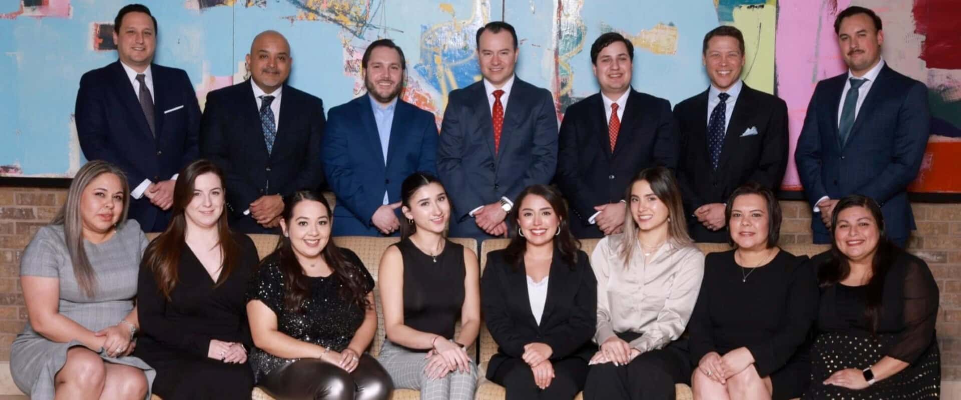 A group photo of 15 people, dressed in business attire, sitting and standing in two rows against a colorful abstract background.
