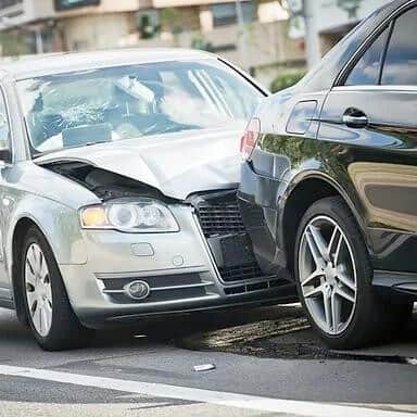 A silver car is rear-ended by a black car on a city street. Both vehicles show damage.