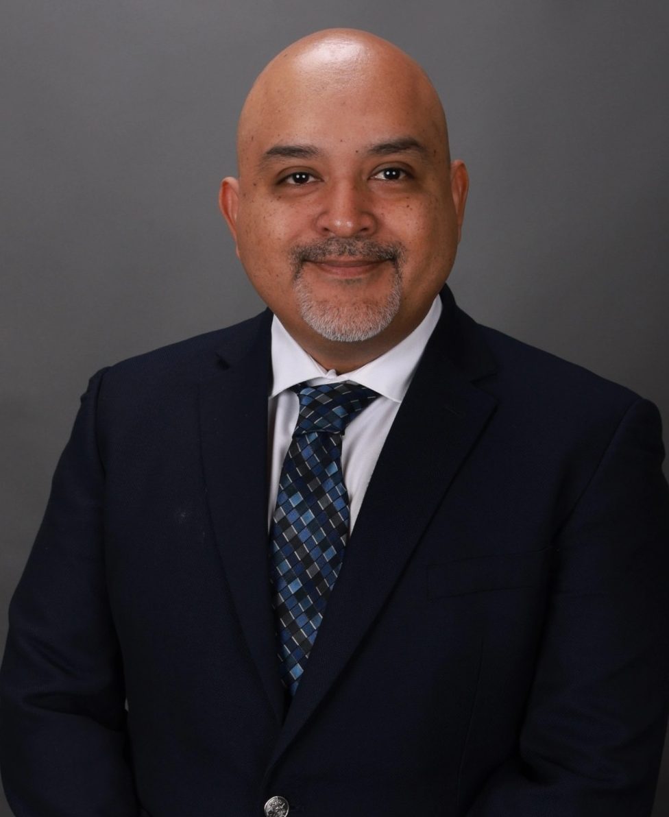 Alberto Rodriguez, Attorney at Law. A person wearing a suit and tie standing against a plain dark background.