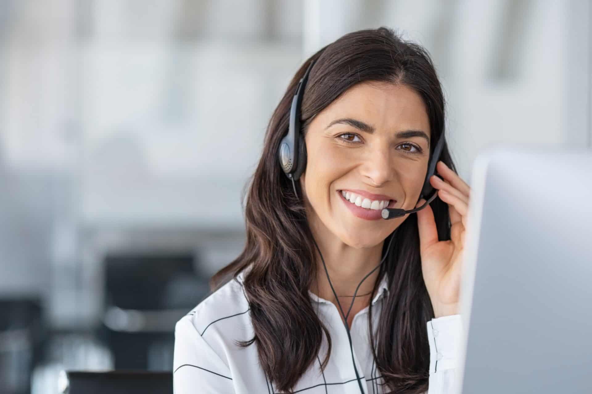 A woman with long dark hair is smiling and wearing a headset in an office setting. She is looking at a computer screen.